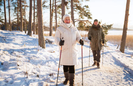 Two people wearing winter coats and walking on a snowy trail with hiking sticks