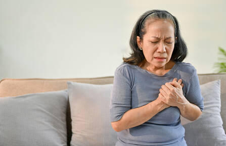 A woman sitting on a sofa with her eyes closed, a pained expression on her face, and her hands over her heart