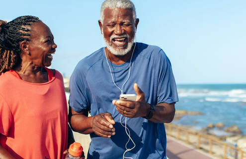 Two people laughing while walking on the beach. One person is looking at their phone and has earbuds in their ears.