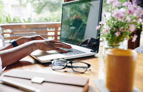 A person working on a laptop computer on a porch table with eye glasses and a vase of flowers