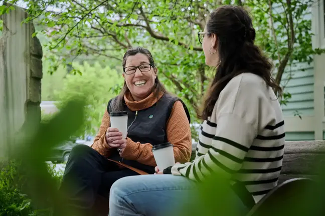 Two women sit on a park bench talking and drinking coffee